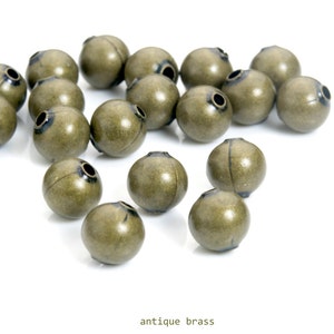 May include: A collection of antique brass beads with a spherical shape and a small hole at the top. The beads have a weathered, aged appearance, and are scattered on a white surface. The text "antique brass" is at the bottom.