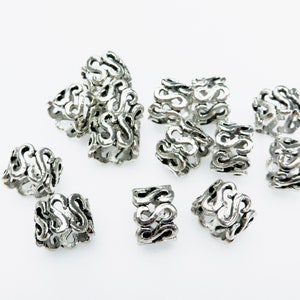 Puede incluir: Un conjunto de cuentas para el cabello de color plateado con un diseño decorativo e intrincado. Las cuentas tienen forma de anillo y presentan un patrón repetitivo de formas estilizadas en forma de 'S'.