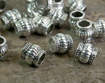 May include: A group of silver metal beads with a decorative design. The beads have a round shape with a raised, textured pattern. The beads are scattered on a textured surface.