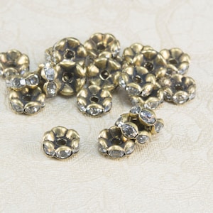 May include: A group of antique gold-toned metal flower-shaped beads with clear rhinestones. The beads are scattered on a white surface.