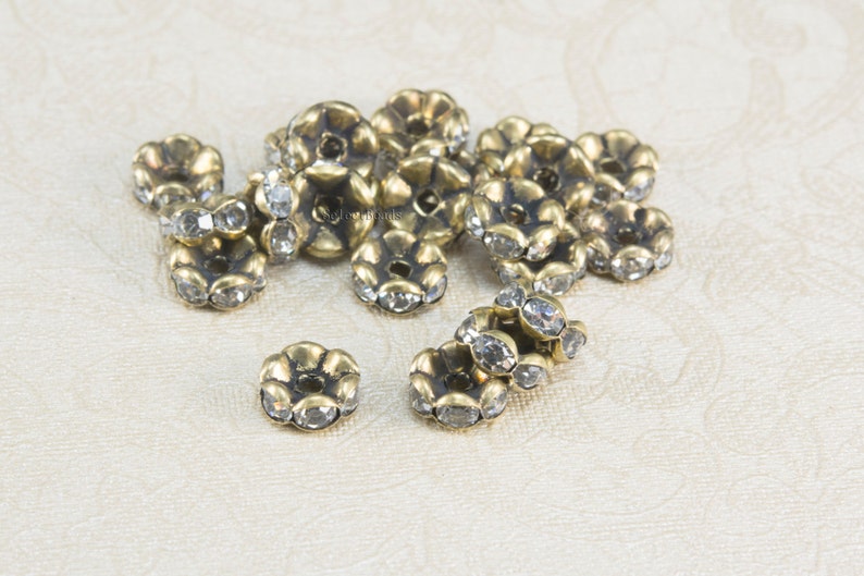 May include: A group of antique gold-toned metal flower-shaped beads with clear rhinestones. The beads are scattered on a white surface.