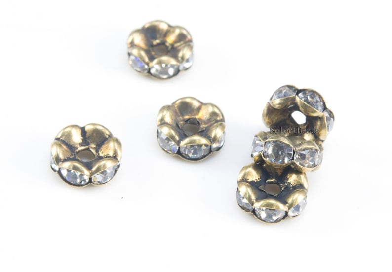 May include: Four antique gold-toned metal spacer beads with clear rhinestone accents. The beads are round with a flower-shaped design.