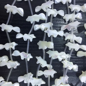 May include: A collection of white, iridescent, shell-shaped pendants, each with a unique design, strung on clear cords. The pendants are arranged in a random pattern, creating a visually appealing display.