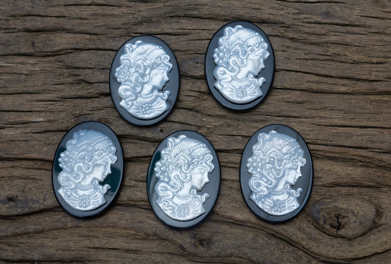May include: Five black cameo pendants with silver-toned raised images of women's profiles. The pendants are oval-shaped and have a smooth, glossy finish.