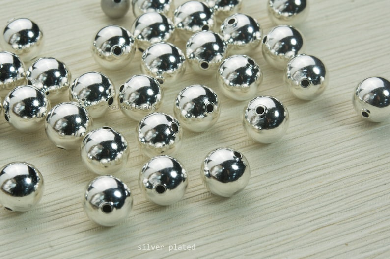 May include: A collection of shiny, silver-plated beads. Each bead is spherical with a smooth, reflective surface and two small holes. The beads are scattered, with the text "silver plated" at the bottom.