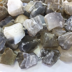 May include: A close-up shot of a collection of rough-cut gemstone beads in shades of grey, white, and tan. The beads are strung together with silver-coloured string, showcasing their natural, irregular shapes. Suitable for jewellery making.
