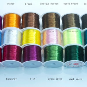 Pode incluir: Bobinas de cordão elástico colorido em várias tonalidades, incluindo roxo, laranja, castanho e preto. A imagem mostra tons rotulados como "fúcsia", "bordô" e "verde relva". Ideal para artesanato e criação de joias.