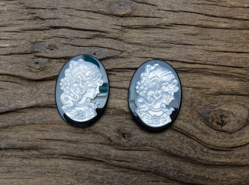May include: Two black cameo pendants with silver-toned raised images of women's profiles. The pendants are oval shaped and have a smooth, glossy finish.