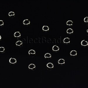 May include: A close-up of a group of silver jump rings scattered on a black surface. The jump rings are small and round, and they are perfect for jewelry making.