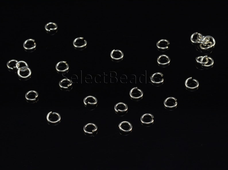 May include: A close-up of a group of silver jump rings scattered on a black surface. The jump rings are small and round, and they are perfect for jewelry making.