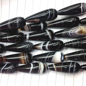 May include: A strand of black and white agate gemstone beads in a teardrop shape. The beads have a polished finish and are strung together on a clear thread.