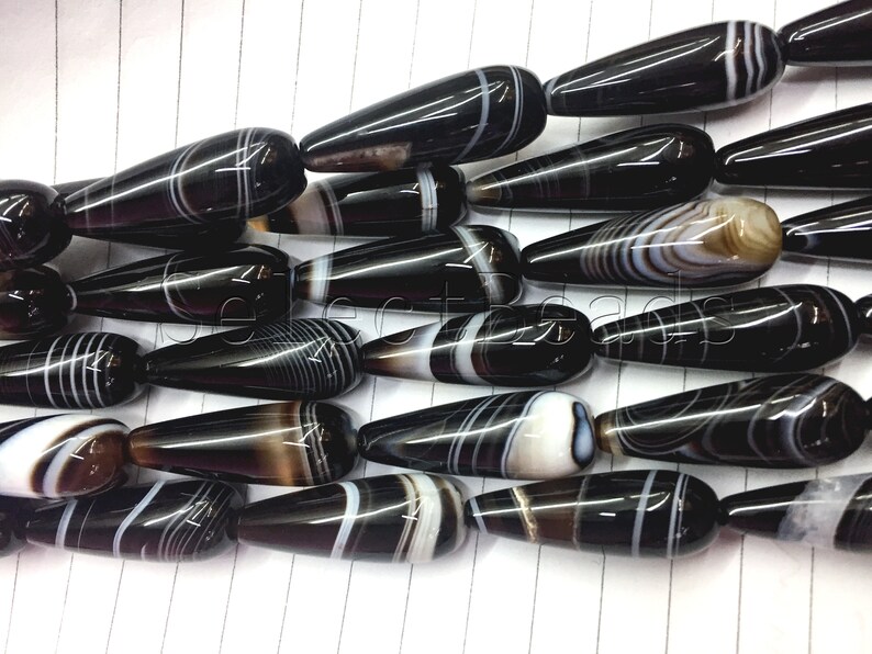 May include: A strand of black and white agate gemstone beads in a teardrop shape. The beads have a polished finish and are strung together on a clear thread.