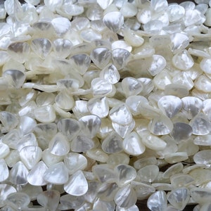 May include: A close-up of a pile of white, iridescent, shell-shaped beads. The beads are all the same size and shape, and they are arranged in a random pattern.