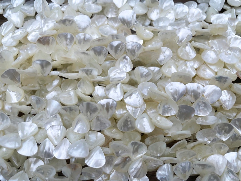 May include: A close-up of a pile of white, iridescent, shell-shaped beads. The beads are all the same size and shape, and they are arranged in a random pattern.