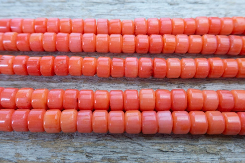 Puede incluir: Una hebra de cuentas de coral, de color naranja brillante, dispuestas en tres filas sobre una superficie de madera.