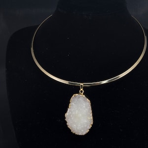 Puede incluir: Un collar choker dorado con un gran colgante de cristal blanco. El colgante es un cristal natural con una superficie rugosa y texturizada.