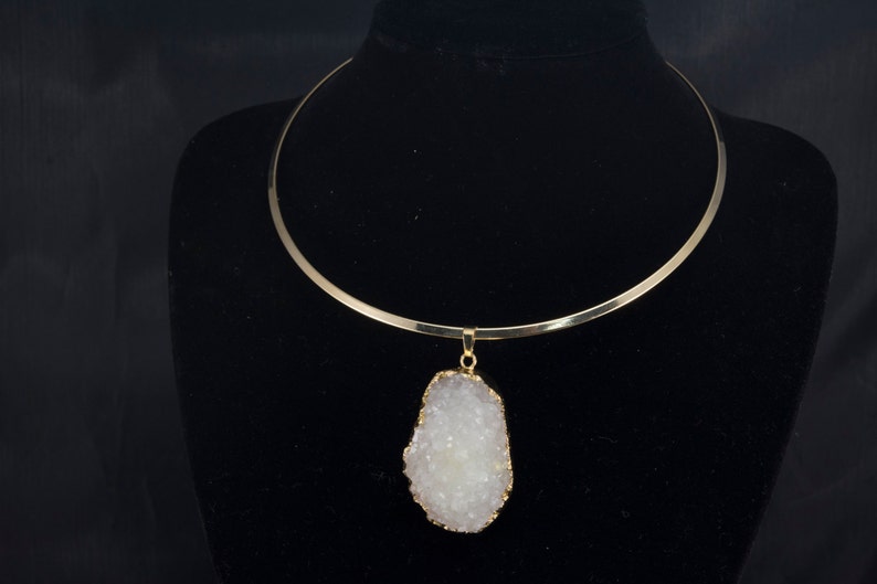 Puede incluir: Un collar choker dorado con un gran colgante de cristal blanco. El colgante es un cristal natural con una superficie rugosa y texturizada.
