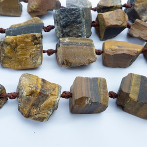 May include: A strand of tiger's eye beads, each bead is a natural, rough-cut, brown and gold stone with dark brown stripes. The beads are strung on a brown cord.