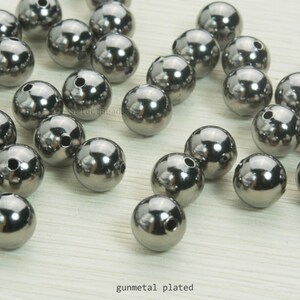 May include: A collection of gunmetal-plated, spherical beads with a reflective surface. Each bead has a small hole drilled through it. The beads are scattered across a light-colored surface. The text "gunmetal plated" is visible.