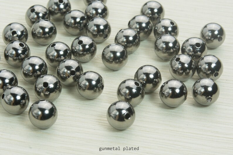 May include: A collection of gunmetal-plated, spherical beads with a reflective surface. Each bead has a small hole drilled through it. The beads are scattered across a light-colored surface. The text "gunmetal plated" is visible.