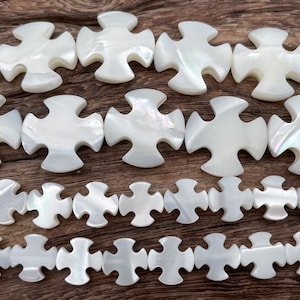 May include: White mother-of-pearl beads in a cross shape with a jigsaw piece design. The beads are strung together in a row.