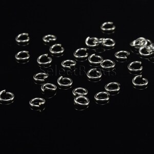 May include: A close-up of a group of silver jump rings scattered on a black surface. The rings are small and round, with a smooth finish.