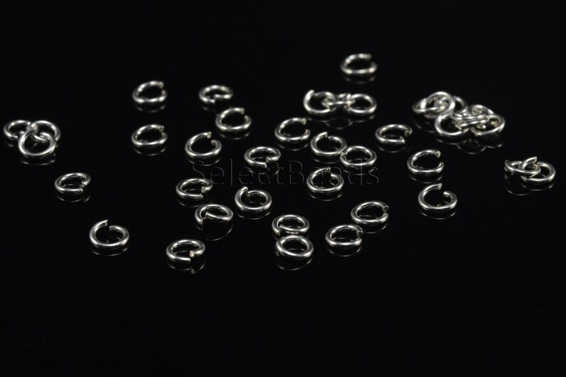 May include: A close-up of a group of silver jump rings scattered on a black surface. The rings are small and round, with a smooth finish.