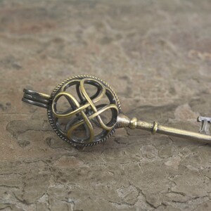 May include: A gold and silver key charm with a decorative, intricate knot design. The key charm is a decorative piece and does not have a functional keyhole.