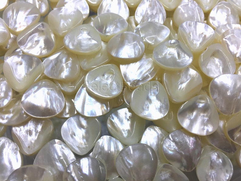 May include: A close-up of a pile of white, iridescent, shell-shaped beads. The beads have a smooth, polished finish and a slightly irregular shape.
