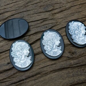 May include: Four oval-shaped black cameo pendants with white raised designs of women's profiles. The pendants are on a wooden surface.