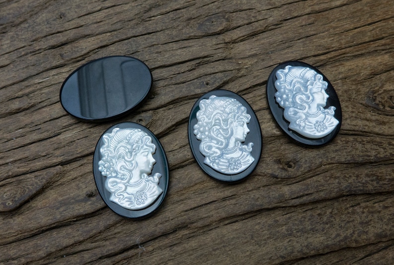 May include: Four oval-shaped black cameo pendants with white raised designs of women's profiles. The pendants are on a wooden surface.