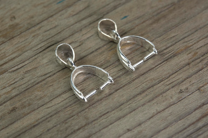 May include: Two silver-toned pendant bails with a loop at the top and a curved opening at the bottom. The bails are designed to hold a pendant or charm.