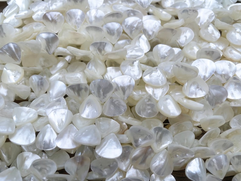 May include: A close-up of a pile of white, pearlescent, triangular-shaped beads. The beads are smooth and shiny, and they have a slightly iridescent sheen.