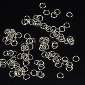 May include: A large quantity of silver jump rings, scattered on a black background. The rings are small and circular, with a smooth, polished finish.
