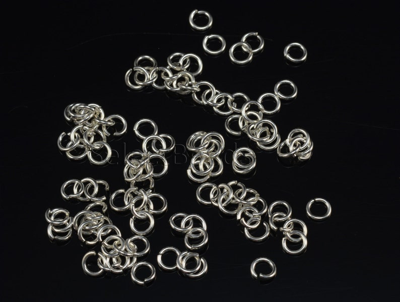 May include: A large quantity of silver jump rings, scattered on a black background. The rings are small and circular, with a smooth, polished finish.