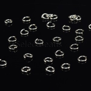 May include: A close-up of a group of silver jump rings scattered on a black surface. The jump rings are small and circular, with a gap in the ring.