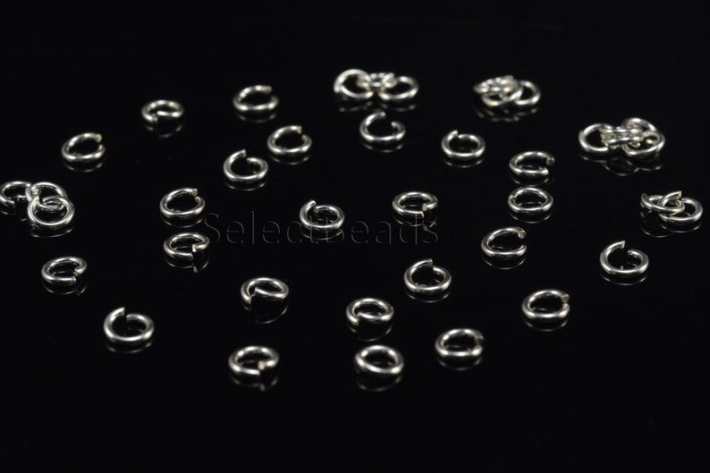 May include: A close-up of a group of silver jump rings scattered on a black surface. The jump rings are small and circular, with a gap in the ring.