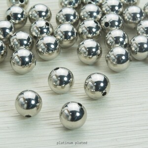 May include: A collection of shiny, platinum-plated, spherical beads. Each bead has a small hole drilled through it. The beads are scattered on a light-colored surface. The text "platinum plated" is visible at the bottom of the image.