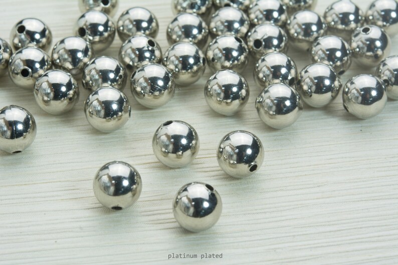 May include: A collection of shiny, platinum-plated, spherical beads. Each bead has a small hole drilled through it. The beads are scattered on a light-colored surface. The text "platinum plated" is visible at the bottom of the image.