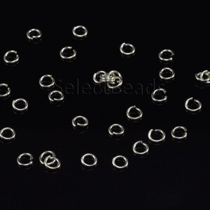 May include: A close-up of a group of silver jump rings, scattered on a black surface. The rings are all the same size and shape, and they are arranged in a random pattern.