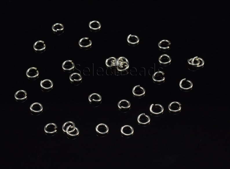 May include: A close-up of a group of silver jump rings, scattered on a black surface. The rings are all the same size and shape, and they are arranged in a random pattern.