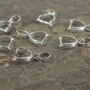 May include: Eight silver-toned pendant bail clasps, each with a loop at the top and a curved, open design at the bottom. The clasps are arranged on a textured, brown surface.