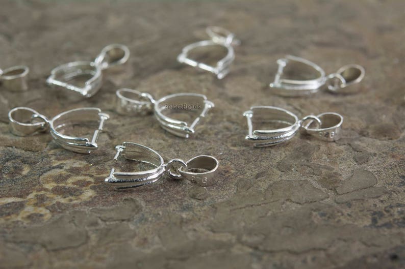 May include: Eight silver-toned pendant bail clasps, each with a loop at the top and a curved, open design at the bottom. The clasps are arranged on a textured, brown surface.