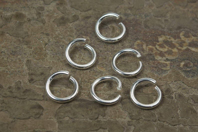 May include: Seven silver jump rings, each with a circular shape and a slightly open gap, are arranged on a textured brown surface.
