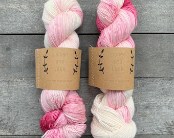 Puede incluir: Dos madejas de hilo teñido a mano con una paleta de colores variados en crema, rosa suave y magenta intenso. Cada madeja está envuelta en una etiqueta de papel marrón impresa con el texto Lichen and Lace y un gráfico de rama de hoja.