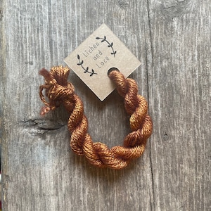 May include: A skein of orange and gold-colored yarn is displayed on a wooden surface. A small, square, brown tag with the text "Lichen and Lace" and a leaf design is attached to the yarn. The yarn is twisted into a circular shape.