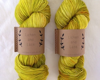 Puede incluir: Dos ovillos de hilo teñido a mano en un color amarillo verdoso brillante con una etiqueta marrón que dice "Lichen and Lace" y "hand dyed in Sackville, NB".