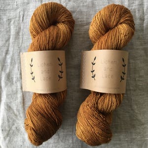 May include: Two skeins of golden-brown yarn with paper labels. The labels read "Lichen and Lace" with leaf illustrations. The yarn is tightly wound, showing texture and color variation. The background is a neutral, light-colored fabric.