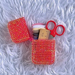 May include: An open, bedazzled, orange-red case, possibly for sewing supplies, with a small ruler, red scissors, and spools of thread. The case is covered in small, sparkling rhinestones. The background is a fluffy, white, textured surface.