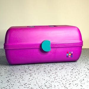 May include: A magenta plastic Caboodles storage case with a rounded rectangular shape, a teal latch on the front, and a teal handle on top. The front features a small, colorful logo with geometric shapes and the text Caboodles printed in small letters.
